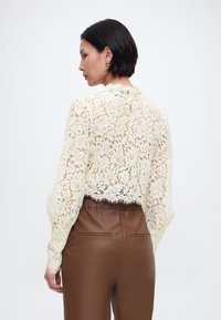 Γυναίκα με κοντά σκούρα μαλλιά, φοράει ανοιχτόχρωμη μπλούζα δαντέλας με μακριά μανίκια και καφέ δερμάτινο παντελόνι ψηλής μέσης, στραμμένη με την πλάτη στην κάμερα.