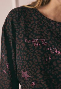 Top in tessuto nero con macchie leopardate marroni, con testo grafico rosa e simboli, tra cui cuori e stelle, in un design rilassato.