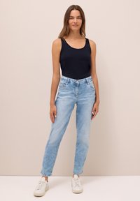 Donna in piedi contro uno sfondo neutro, indossa una canottiera nera, jeans azzurri chiari e sneakers bianche, con le mani rilassate ai lati.