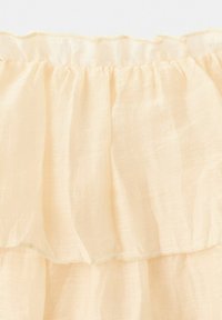 Světlé béžové průsvitné látky s nařasenými volánovými vrstvami a jemnými šicími detaily na okrajích.