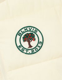 Krēmkrāsas pūkains jakas ar apļa izšuvumu, kurā attēlots koks, sarkanie āboli un uzraksts "BLKVIS EST. 2013" zaļā krāsā.
