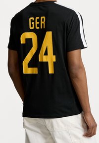 Camiseta negra con el texto amarillo "GER" y "24" en la espalda, con rayas laterales blancas, hecha de algodón y de manga corta.