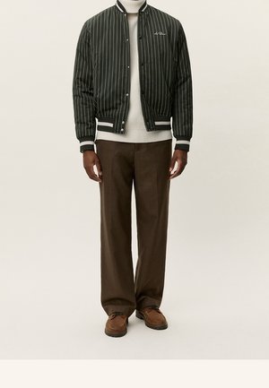 Man wearing black pinstripe bomber jacket, white turtleneck sweater, brown trousers, and brown casual shoes standing against plain background.