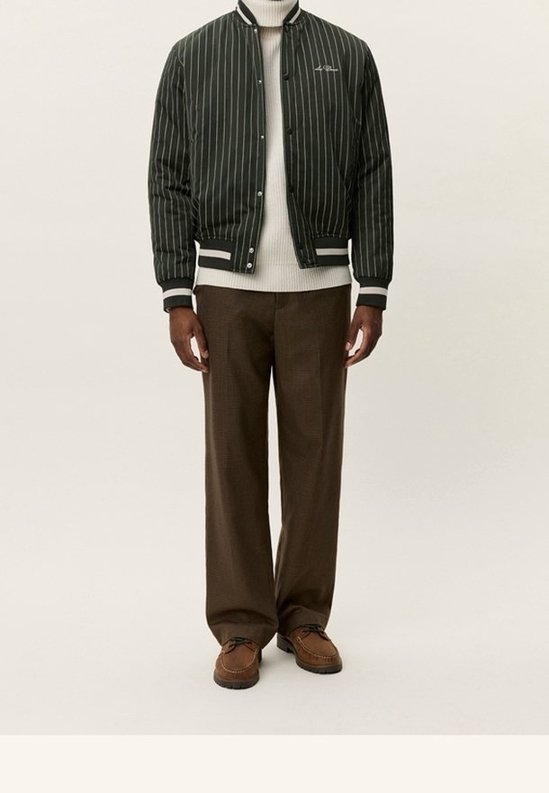Homme portant une veste bomber noire à fines rayures, un pull à col roulé blanc, un pantalon marron et des chaussures décontractées marron, debout devant un fond uni.