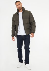 Olivengrøn pufferjakke med høj krave, med lynlås foran og to sidelommer, parret med mørk denimjeans og hvide sneakers.