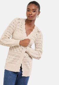 Könnyű bézs kötött kardigán, bonyolult mintákkal, V-nyakkal, gombos záródással, valamint bordázott ujjal és aljjal. Farmer dzseki fölé viselve.