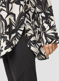 Vloeiende blouse met een zwart-wit bloemenpatroon en gouden accenten, afgeronde zoom en losse mouwen, gecombineerd met zwarte broeken.