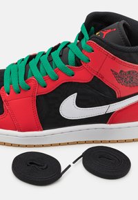 Černé a červené tenisky Air Jordan s zelenými šňůrkami a bílou podrážkou, doplněné dvěma extra zkroucenými černými tkaničkami na bílém pozadí.