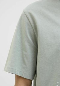 Camiseta de algodón verde claro con un ajuste relajado, mangas cortas y cuello redondo. Textura suave con detalles de costura sutiles cerca del dobladillo.
