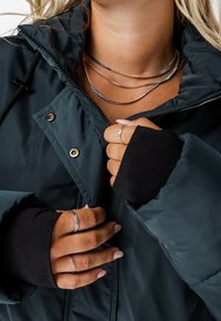 Gevoerde donkerblauwe jas met rits en drukknoopsluiting, met zwarte duimgaten in de manchet. Gelaagde zilveren halskettingen en ringen op de handen.
