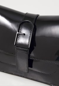 Sac à main noir brillant avec une finition en vernis lisse. Dispose d'une fermeture type ceinture avec une boucle noire brillante et des détails cousus.