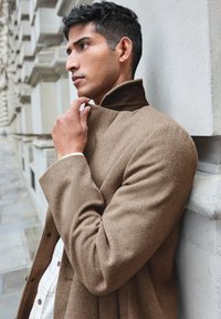 Cappotto di lana marrone con un colletto alto, in tessuto texturizzato. Il colletto interno è di un marrone scuro, sovrapposto a una camicia a quadri bianchi.