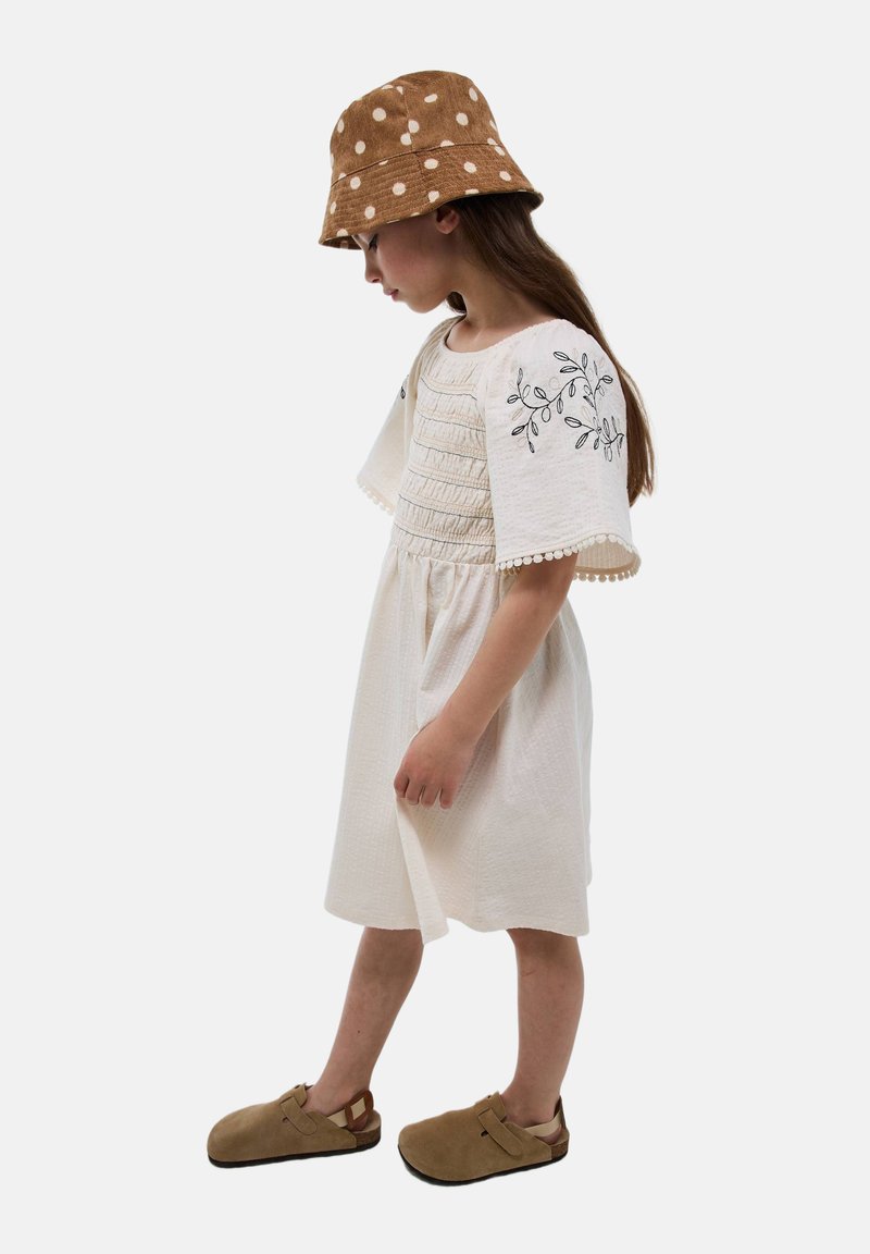 Robe blanche brodée avec des manches évasées et un corsage froncé, associée à un chapeau seau à pois marron et des sabots beiges.