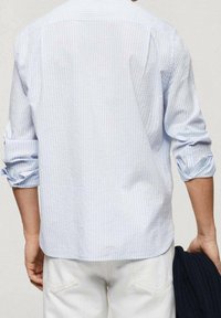 Světle modrá pruhovaná košile s dlouhými rukávy, měkká tkanina, uvolněný střih a zakřivený lem. Nosí se s bílými kalhotami, s vyhrnutými rukávy.