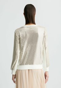 Maglione dorato con paillettes, maniche lunghe, polsini bianchi e collo a giro. Texture liscia con una superficie lucida e riflettente.