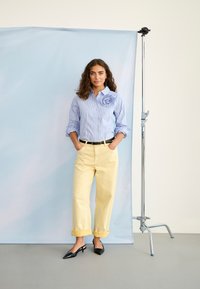 Žena s kudrnatými vlasmi v modrej prúžkovanej košeli s kvetinovým vzorom, žltými vyhrnutými nohavicami a čiernymi topánkami so zádierkami stojí pri stojane s pozadím.