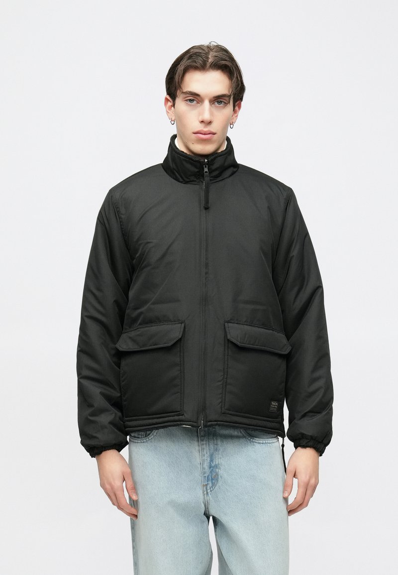 Casaco acolchoado preto com gola alta, fecho frontal de correr e dois grandes bolsos. Punhos e bainha elásticos com textura de tecido suave e mate.