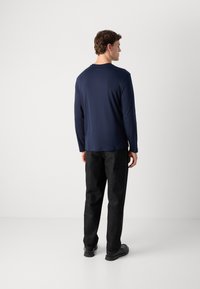 Man wearing a navy long-sleeve shirt, black pants, and black shoes, standing facing away against a plain white background.