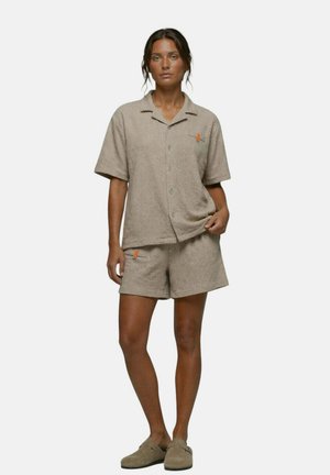 Femme debout portant une chemise beige texturée à manches courtes boutonnée et un short assorti avec des chaussures beiges décontractées sans lacets.