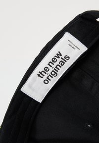Svart stofflue med en hvit etikett som står "the new originals." Har en buet skygge og broderte detaljer. Én størrelse.