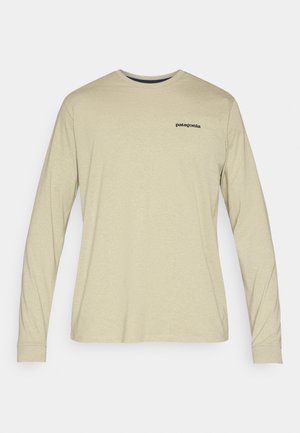 Košile s dlouhým rukávem z lehké béžové látky s kulatým výstřihem. Na horní levé straně je malá černá logo "Patagonia". Hladká textura.