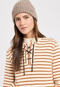 Beige og brunstribet top med snørret hals, flæset krave og en strikket brun hue båret over skulderlangt hår.
