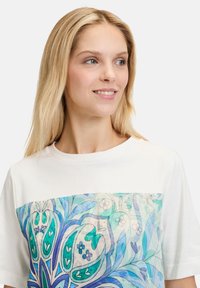 Hvid bomulds T-shirt med et stort blåt og grønt blomstermotiv på forsiden, rund halsudskæring og korte ærmer.