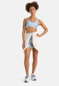 Reggiseno sportivo blu con motivo e pantaloni shorts abbinati, con una maglietta crema annodata intorno alla vita. Sneakers bianche con accenti blu.