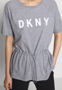 Camiseta de algodón gris con mangas cortas de raglán, que presenta un logo blanco "DKNY", cinturilla con cordón y bolsillos laterales. Textura suave.