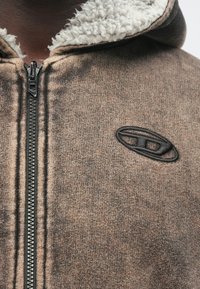 Nahaufnahme eines braunen Kapuzenpullovers mit Reißverschluss, Fleece-Futter und einem schwarzen, aufgestickten, ovalen Logo auf der Brust.