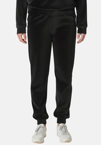 Zwarte polyester joggingbroek met een gladde textuur, elastische tailleband en taps toelopende pijpen, gecombineerd met witte sneakers. Geen patronen of accenten zichtbaar.