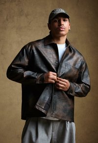 Barna bőrkabát laza fazonban, gombos záródással, zsebekkel és enyhén fényes textúrával. Fehér ing felett viselve.