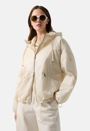 Woman wearing oversized cream puffer jacket with hood, cream pants, and white round sunglasses, standing with one hand in pocket.