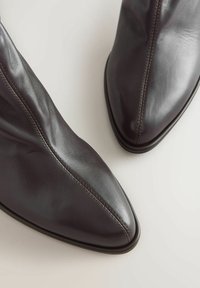 Tumed pruunid nahast saapad terava nina ja nähtava õmblusega keskelt ees, nähtud ülevalt kerge tausta peal.