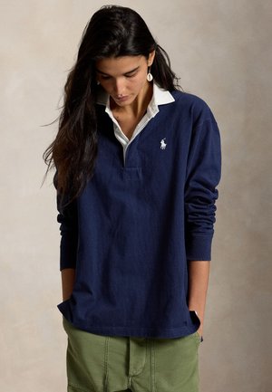 Femme aux longs cheveux foncés portant un maillot de rugby bleu marine avec un col blanc, un pantalon vert et des boucles d'oreilles en coquillage, regardant vers le bas.