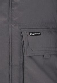 Szürke szigetelt dzseki texturált anyagból. Elöl zsebfedős zseb, márkázott címke és varrással díszített szegélyek találhatók rajta.