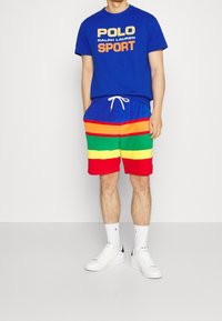 Man som bär en blå Polo Ralph Lauren sport t-shirt och flerfärgade randiga shorts med vita strumpor och vita sneakers, stående mot en vit vägg.