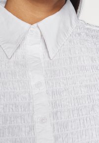 Hvid skjorte med krave, der har tekstureret smock-detalje, med knapfront og glat stof. Knapperne er af klart plast, hvilket forbedrer designet.