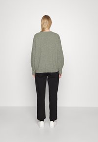 Olivgrüner Strickpullover mit rundem Ausschnitt, überschnittenen Schultern und gerippten Bündchen, kombiniert mit schwarzer Hose und weißen Sneakern.