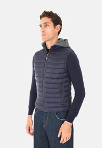 Giovane uomo che indossa un gilet trapuntato blu navy con cappuccio grigio su una camicia a maniche lunghe blu navy, in piedi contro uno sfondo chiaro e uniforme.