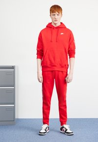 Rdeča kapuca s kapuco in kenguru žepom, v kombinaciji z usklajenimi trenirkami, ki imajo bele stranske črte. Črno-bele superge.