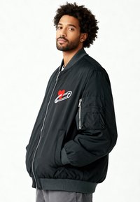 Veste bomber noire avec col et poignets côtelés. Présente un motif de cœur rouge et une épingle de sûreté sur la poitrine. Poches latérales et poche zippée sur la manche.