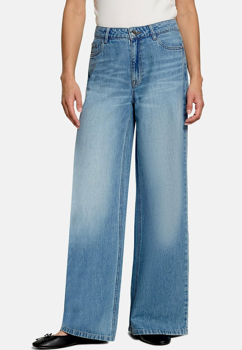 Femme portant un jean large bleu clair avec des coutures visibles et des chaussures plates noires, debout devant un fond uni.