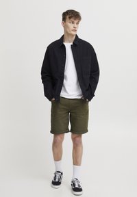 Cămașă neagră cu butoni, tricou alb, pantaloni scurți verde măsliniu cu umăr răsuciți, șosete albe până la glezne, adidași negri. Design simplu și aspect casual.