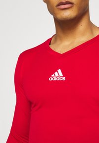 Rode sportieve shirt met lange mouwen en V-hals, met een textuurstof en een wit Adidas-logo op de linkerborst.