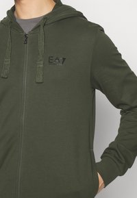 Felpa con cappuccio verde con cerniera, realizzata in tessuto morbido, con cappuccio, logo "EA7" in rilievo e coulisse testurizzate.