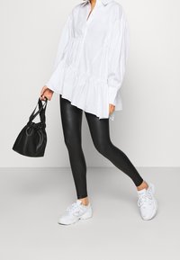 Žena oblečená v oversize bílé košili s knoflíky, černých kožených legínách, bílých teniskách a držící malou černou kabelku na bílém pozadí.