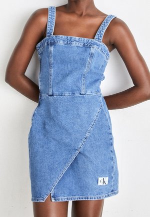 Femme portant une mini robe en denim bleu clair avec larges bretelles, encolure carrée, et jupe portefeuille asymétrique avec un petit écusson logo.