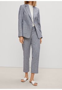 Completo grigio chiaro in tessuto testurizzato, giacca su misura con rever a punta, pantaloni coordinati e sandali bianchi senza lacci.