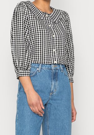 Frau trägt eine schwarz-weiß karierte Bluse mit Puffärmeln und blaue, hoch taillierte Jeans, steht mit einer Hand herunter hängend.